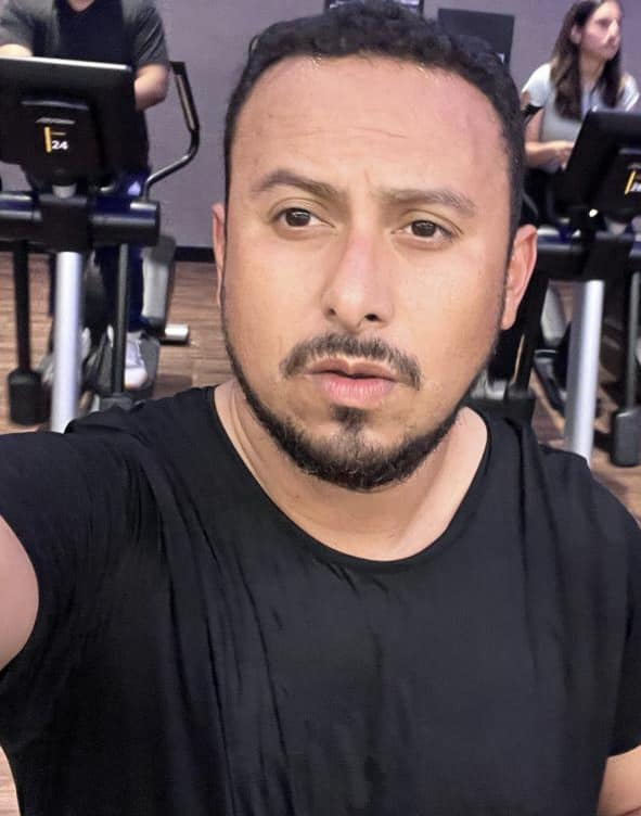 Selfie de Servando Salazar Cano en el gimnasio, vistiendo una camiseta negra y con una expresión seria. Se pueden ver personas ejercitándose en el fondo.